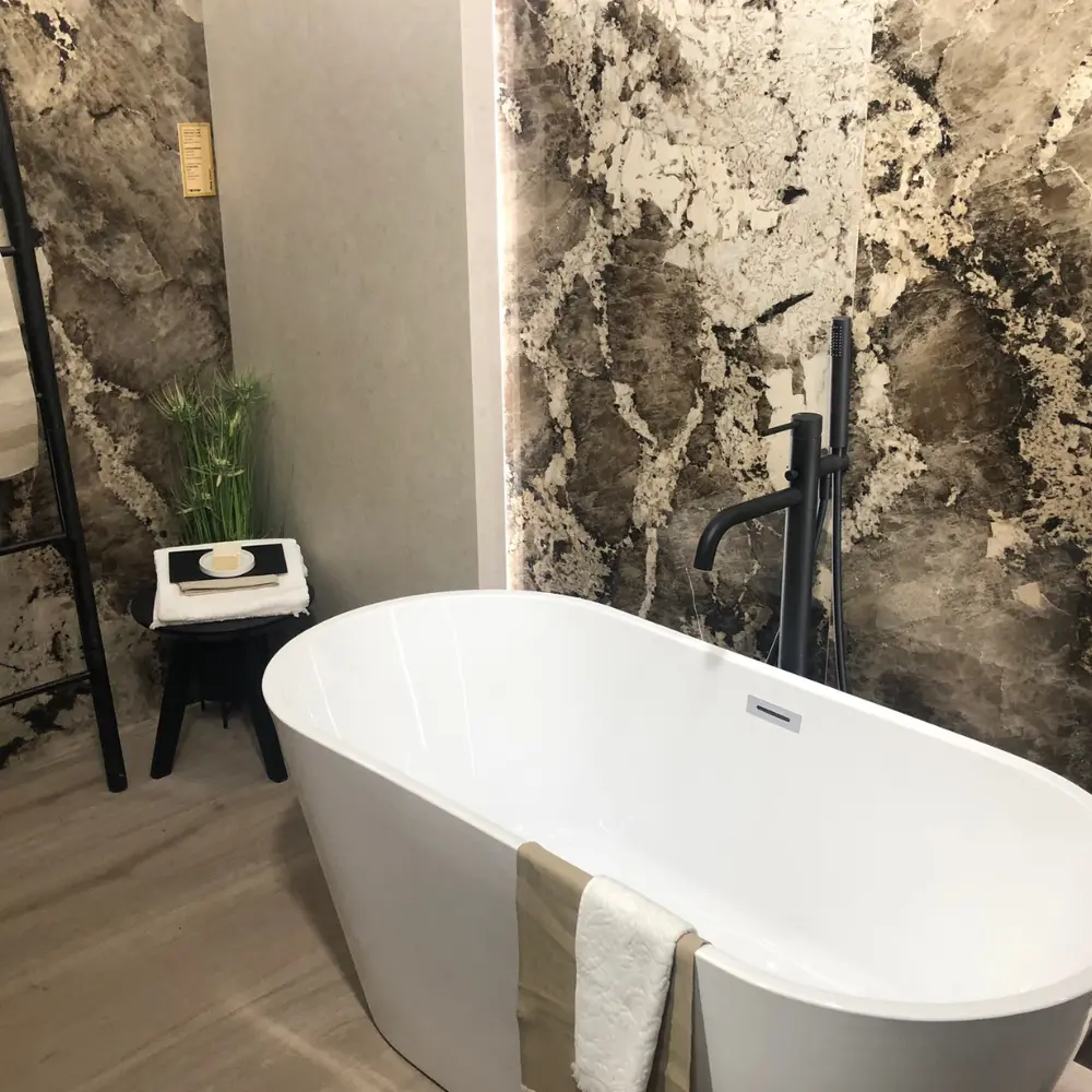 Freistehende weiße Badewanne mit schwarzer Standarmatur vor einer großformatigen Wandfliese in dunkler Marmoroptik mit markanter Maserung, hinterleuchtetem Wandpaneel und schwarzem Beistellhocker – Baddesign aus Korschenbroich