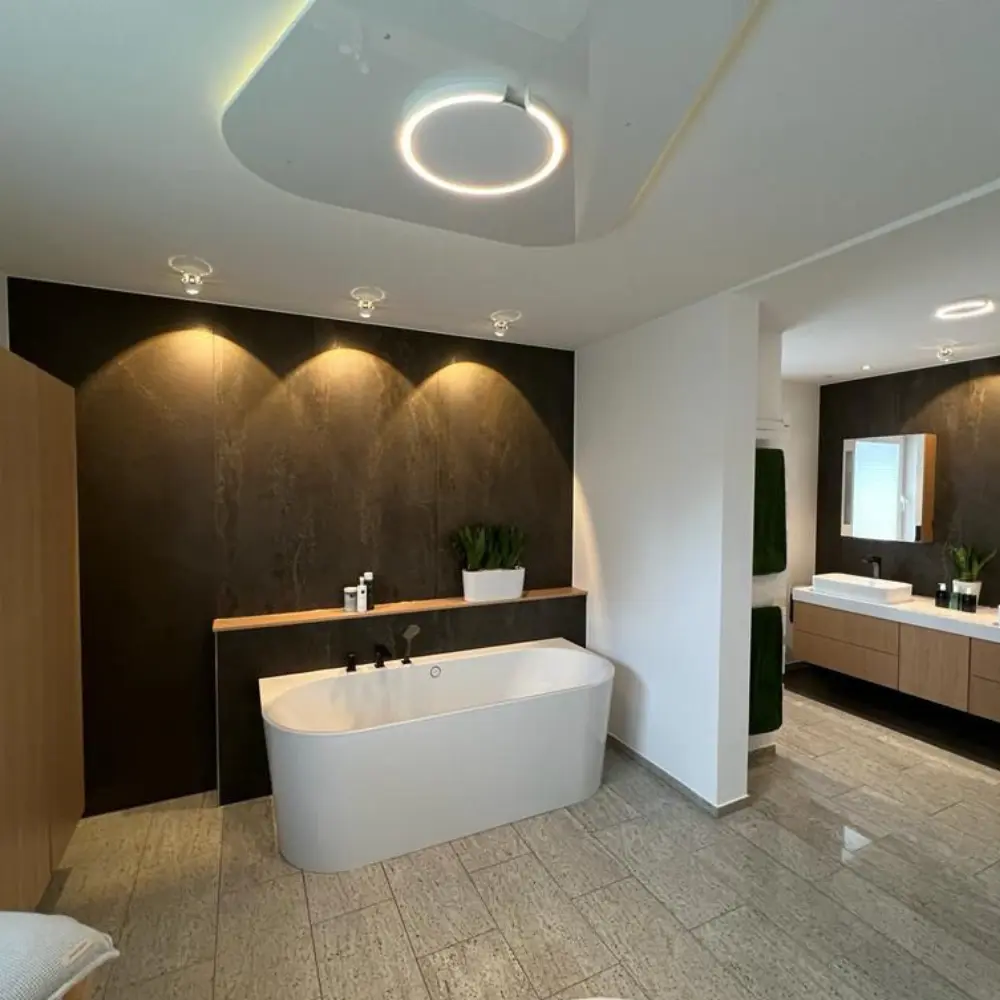 Elegantes Badezimmer mit freistehender Badewanne vor einer dunklen XXL-Keramik-Wandverkleidung in Natursteinoptik, warmem Akzentlicht, Eichenholz-Waschtisch und hellem Steinboden – realisiert in Korschenbroich bei Düsseldorf