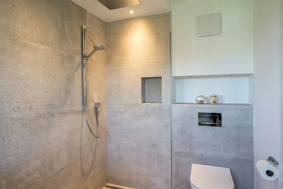 Kompaktes modernes Badezimmer mit bodengleicher Walk-In-Dusche, großformatigen Wandfliesen in heller Betonoptik, Regenduschkopf in der Decke, gefliester Wandnische und wandhängendem WC – Fliesenverlegung aus Korschenbroich